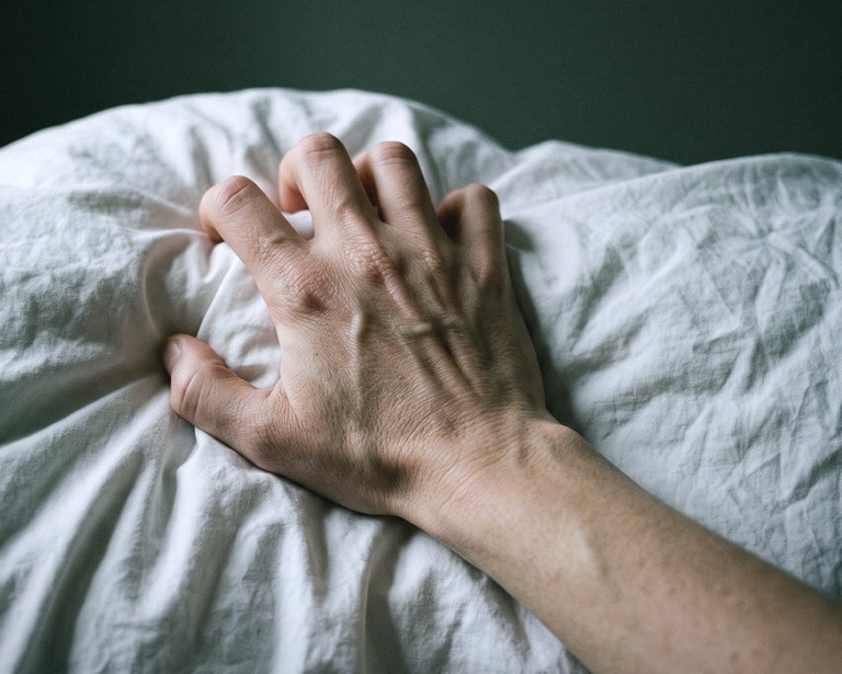 Mão de uma mulher apertando o lençol durante a mamada, tendões e nós dos dedos tensionados, em luz fria de madrugada.